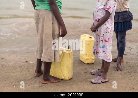 Uganda - November, 26 2016: Beschnittene, nicht erkennbare kleine afrikanische Mädchen, die schwere Kanister mit Wasser aus dem Fluss tragen und an der nassen Küste in der Nähe von Mitkindern im Dorf spazieren gehen Stockfoto