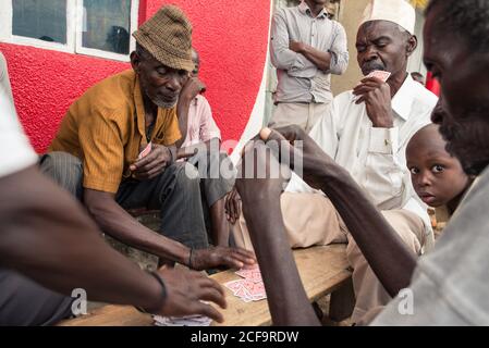 Uganda - November, 26 2016: Gruppe von schwarzen Männern in legerer Kleidung spielen Kartenspiel, während sie um die Bank auf der Straße der Stadt sitzen Stockfoto