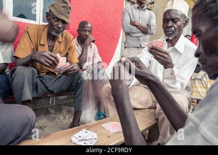 Uganda - November, 26 2016: Gruppe von schwarzen Männern in legerer Kleidung spielen Kartenspiel, während sie um die Bank auf der Straße der Stadt sitzen Stockfoto