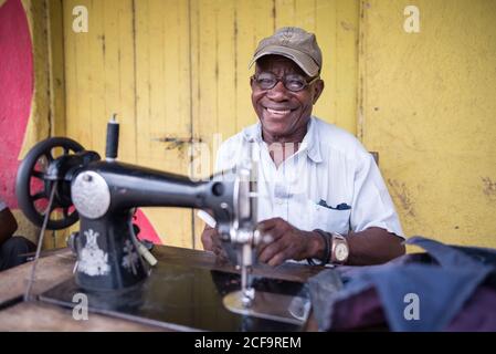 Uganda - November, 26 2016: Glücklicher schwarzer Mann in Hemd und Mütze lächelnd und beim Blick auf die Kamera, während er die Nähmaschine vor der schäbigen Werkstatt auf der Straße benutzt Stockfoto