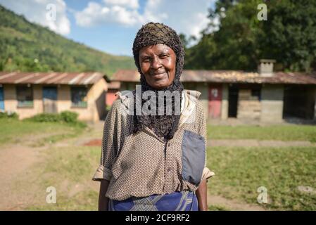 Uganda - November, 26 2016: Freundliche reife schwarze Frau im Kopftuch lächelnd und mit Blick auf die Kamera, während sie an sonnigen Tagen im Dorf gegen Häuser steht Stockfoto