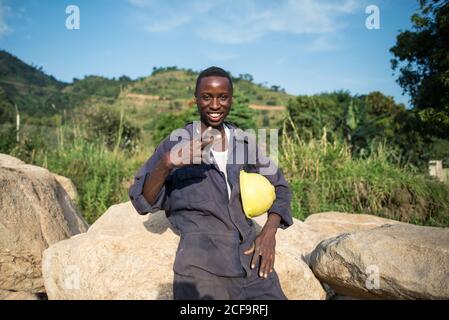 Uganda - November, 26 2016: Glücklicher schwarzer Baumeister mit Hardhat lächelnd und gestikulierenden V-Zeichen, während er sich auf Steinen gegen Hügel in der Landschaft lehnt Stockfoto