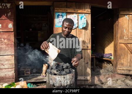 Uganda - 26. November 2016: Niedriger Winkel des ruhigen erwachsenen afrikanischen Mannes, der Papier verbrennt, der in Brazier Holzkohle macht, während er an sonnigen Tagen auf der Straße vor dem Eingang eines kleinen rustikalen Holzgebäudes steht Stockfoto