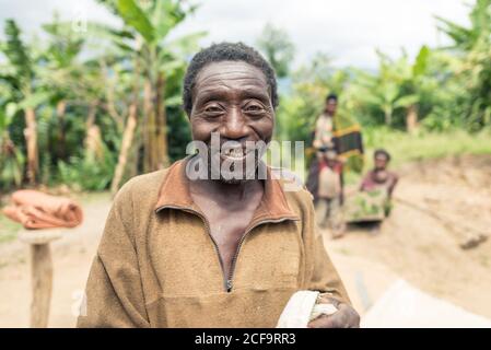 Uganda - November, 26 2016: Älterer Mann, der bei Plantagen gegen grüne Bäume steht Stockfoto