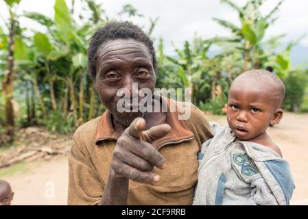 Uganda - November, 26 2016: Älterer Mann, der während der Plantage gegen grüne Bäume steht und ein Enkelkind trägt Stockfoto