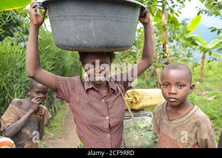 Uganda - November, 26 2016: Afrikanisches Mädchen mit Becken auf dem Kopf und Blick auf die Kamera während der Plantage in der Nähe von schwarzen Freunden Stockfoto