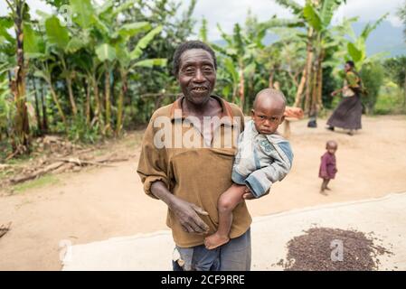 Uganda - November, 26 2016: Älterer Mann, der während der Plantage gegen grüne Bäume steht und ein Enkelkind trägt Stockfoto