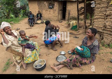 Uganda - November, 26 2016: Schwarze Frauen mit Babys sitzen auf sandigen Boden und kommunizieren miteinander außerhalb schäbiger Hütten auf Dorfstraße Stockfoto