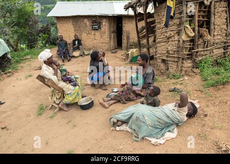 Uganda - November, 26 2016: Schwarze Frauen mit Babys sitzen auf sandigen Boden und kommunizieren miteinander außerhalb schäbiger Hütten auf Dorfstraße Stockfoto