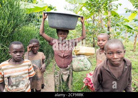 Uganda - November, 26 2016: Afrikanisches Mädchen mit Becken auf dem Kopf und Blick auf die Kamera während der Plantage in der Nähe von schwarzen Freunden Stockfoto
