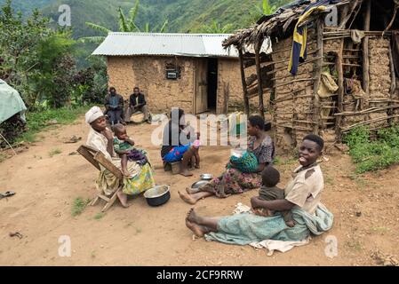 Uganda - November, 26 2016: Schwarze Frauen mit Babys sitzen auf sandigen Boden und kommunizieren miteinander außerhalb schäbiger Hütten auf Dorfstraße Stockfoto