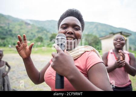 Uganda - 26. November 2016: Fröhliche Afrikanerin in rosa Hemd mit Mikrofon und Gesang, während das Publikum mit Interesse zuhörte und gegen verschwommene grüne Hügel und kleine Gebäude im Dorf klatschte Stockfoto