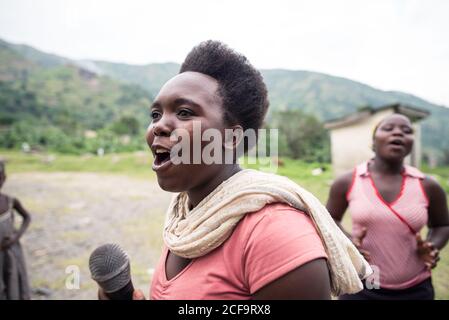 Uganda - 26. November 2016: Fröhliche Afrikanerin in rosa Hemd mit Mikrofon und Gesang, während das Publikum mit Interesse zuhörte und gegen verschwommene grüne Hügel und kleine Gebäude im Dorf klatschte Stockfoto