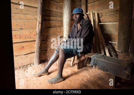 Uganda - 26. November 2016: Friedlicher afrikanischer Barfußmännchen in lässiger Kleidung und blauem Hut, der wegschaut und träumt, während er auf einer Holzbank in einem kleinen dunklen Holzhaus sitzt Stockfoto