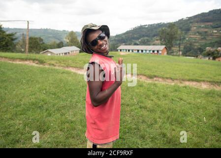 Uganda - November, 26 2016: Freundlicher schwarzer Mann mit Hut und Sonnenbrille, der vor dem Dorf auf Gras steht und die Kamera anschaut Stockfoto