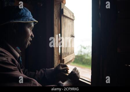 Uganda - 26. November 2016: Fröhlicher erwachsener afrikanischer Rüde in braunem Hemd und blauem Hut, der sich auf ein Fensterloch stützt und die Kamera anlächelt, während er in einem kleinen Holzhaus steht Stockfoto