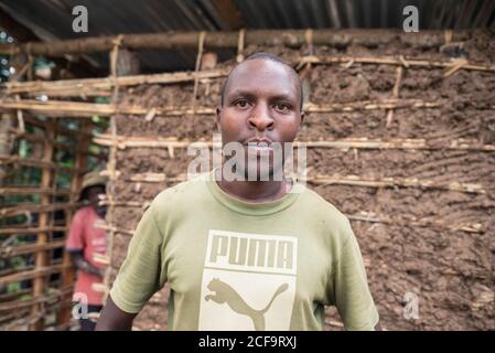 Uganda - 26. November 2016: Ruhiger afrikanischer Mann, der auf die Kamera schaut, die außerhalb des Holzschliff mit braunem Schmutz unter dem Metalldach steht Stockfoto