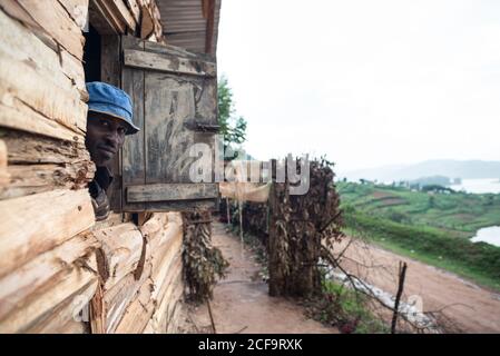 Uganda - 26. November 2016: Fröhlicher erwachsener afrikanischer Rüde in braunem Hemd und blauem Hut, der sich auf ein Fensterloch stützt und die Kamera anlächelt, während er in einem kleinen Holzhaus steht Stockfoto