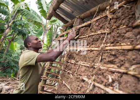 Uganda - 26. November 2016: Seitenansicht eines ruhigen afrikanischen Mannes, der Holzmahlen mit braunem Dreck ausfüllt, um zukünftige Ziegelsteine im Sonnenlicht unter einem Metalldach zu trocknen, während er in einer eigenen Werkstatt vor einem grünen tropischen Wald steht Stockfoto