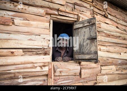 Uganda - 26. November 2016: Fröhlicher erwachsener afrikanischer Rüde in braunem Hemd und blauem Hut, der sich auf ein Fensterloch stützt und die Kamera anlächelt, während er in einem kleinen Holzhaus steht Stockfoto