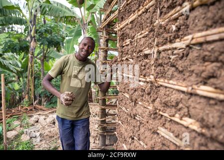 Uganda - 26. November 2016: Seitenansicht eines ruhigen afrikanischen Mannes, der Holzmahlen mit braunem Dreck ausfüllt, um zukünftige Ziegelsteine im Sonnenlicht unter einem Metalldach zu trocknen, während er in einer eigenen Werkstatt vor einem grünen tropischen Wald steht Stockfoto