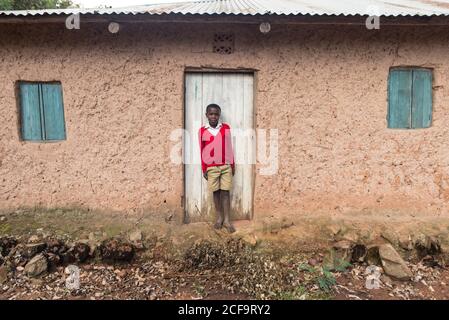 Uganda - November, 26 2016: Afrikanisches Kind, das in einem Dorf auf einer verwitterten Lehmhaustür steht und dabei die Kamera anschaut Stockfoto