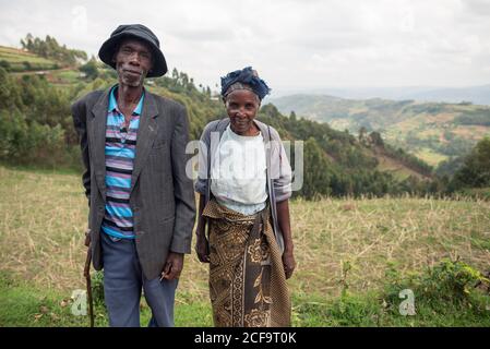 Uganda - 26. November 2016: Nachdenklicher älterer afrikanischer Ehemann mit Frau in legerer Kleidung, der mit freundschaftlicher Sicht vor der erstaunlichen unscharfen Landschaft der grünen bewaldeten Hügel bei Tag die Kamera anschaut Stockfoto