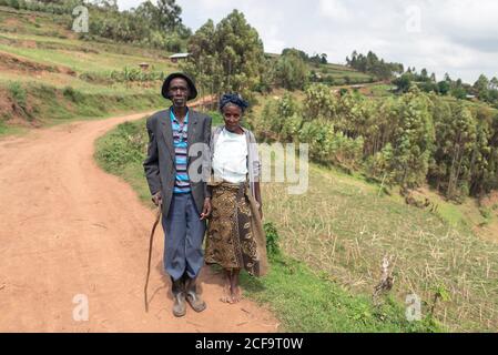 Uganda - 26. November 2016: Nachdenklicher älterer afrikanischer Ehemann mit Frau in legerer Kleidung, der mit freundschaftlicher Sicht vor der erstaunlichen unscharfen Landschaft der grünen bewaldeten Hügel bei Tag die Kamera anschaut Stockfoto