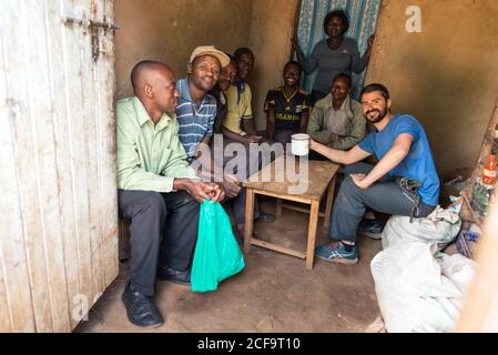 Uganda - 26. November 2016: Fröhlicher Reisender und afrikanischer Ureinwohner lächeln vor der Kamera, während sie am Holztisch sitzen und gemeinsam aus einem allgemeinen Metallbecher trinken Stockfoto