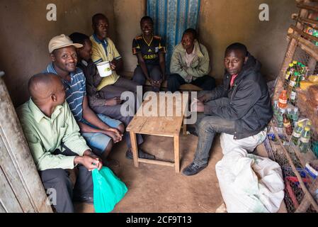 Uganda - 26. November 2016: Fröhlicher Reisender und afrikanischer Ureinwohner lächeln vor der Kamera, während sie am Holztisch sitzen und gemeinsam aus einem allgemeinen Metallbecher trinken Stockfoto