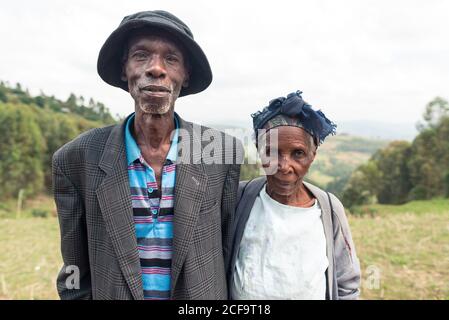 Uganda - 26. November 2016: Nachdenklicher älterer afrikanischer Ehemann mit Frau in legerer Kleidung, der mit freundschaftlicher Sicht vor der erstaunlichen unscharfen Landschaft der grünen bewaldeten Hügel bei Tag die Kamera anschaut Stockfoto