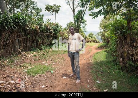 Uganda - November, 26 2016: Freundlicher schwarzer Mann, der auf die Kamera schaut, während er auf der Feldweg-Straße im Dorf steht Stockfoto