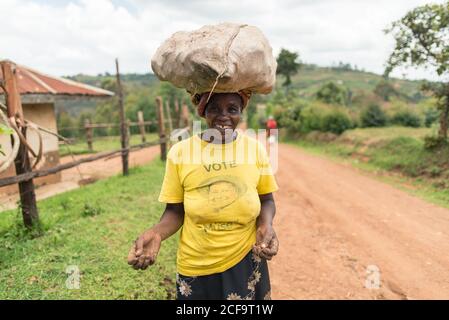 Uganda - November, 26 2016: Ganzkörper Senior African Lady Blick auf Kamera und mit Sack auf den Kopf, während zu Fuß auf Feldweg Stockfoto