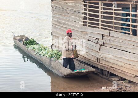 Uganda - 26. November 2016: Ein nachdenklicher afrikanischer Mann, der in einem Holzboot mit grünen Bananen und Kohl gegen ein rustikales Holzgebäude am Pier steht, während er sich darauf vorbereitet, Lebensmittel auf dem lokalen Markt zu verkaufen Stockfoto