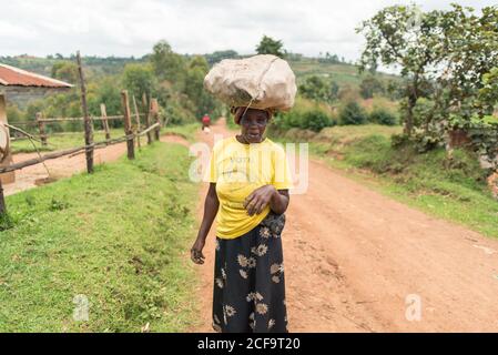 Uganda - November, 26 2016: Ganzkörper Senior African Lady Blick auf Kamera und mit Sack auf den Kopf, während zu Fuß auf Feldweg Stockfoto