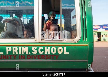 Uganda - 26. November 2016: Aufgeregte afrikanische Kinder in farbenfroher Freizeitkleidung, die neugierig aus dem Fenster schauen und die Kamera anlächeln, während sie im Bus sitzen und an sonnigen Tagen zur Schule gehen Stockfoto