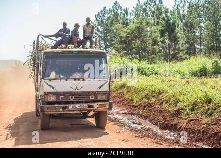 Uganda - 26. November 2016: Aufgeregte afrikanische Arbeiter sitzen auf dem Trailerzaun auf dem Kabinendach und lächeln vor der Kamera, während ein Mitarbeiter an heißen sonnigen Tagen mit einem LKW Fracht auf unbefestigten Straßen transportiert Stockfoto