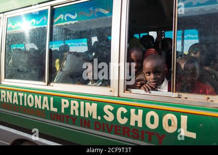 Uganda - 26. November 2016: Aufgeregte afrikanische Kinder in farbenfroher Freizeitkleidung, die neugierig aus dem Fenster schauen und die Kamera anlächeln, während sie im Bus sitzen und an sonnigen Tagen zur Schule gehen Stockfoto