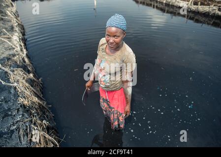 Uganda - 26. November 2016: Nachdenkliche lächelnde afrikanische Frau in lebhafter Freizeitkleidung, die schwarzen salzigen Schlamm konzentriert und abgebaut, während sie im dunklen Wasser steht und Metallwerkzeug auf der Salzfarm benutzt, gegen verschwommene Sicht auf Plantagen und klaren blauen Himmel an heißen Tagen Stockfoto