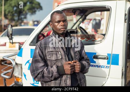 Uganda - November, 26 2016: Freundlicher junger schwarzer Mann, der in der Nähe eines weißen Transporters in der Mitte einer Straße im Dorf auf die Kamera schaut Stockfoto