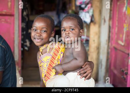 Uganda - November, 26 2016: Afrikanisches Kind, das einen kleinen Bruder hält und vor einem Dorf die Kamera anschaut Stockfoto