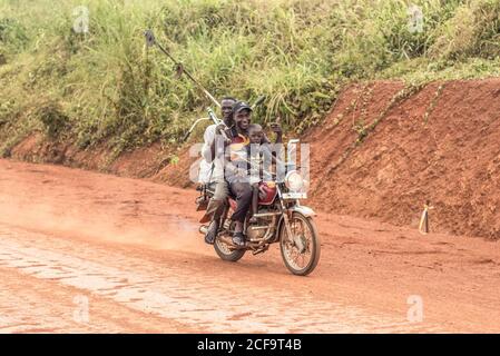 Uganda - 26. November 2016: Aufgeregte afrikanische Arbeiter und kleiner Junge lächeln an der Kamera und geben wie beim Fahren Motorrad auf Feldstraße gehen gegen grüne grasbewachsenen Hügel bei Tageslicht zu arbeiten Stockfoto