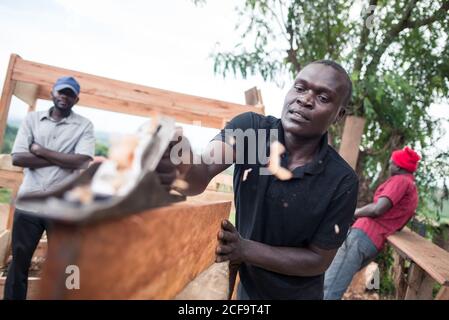 Uganda - 26. November 2016: Niedriger Winkel des fleißigen aufmerksamen afrikanischen Zimmermanns mit Handfläche während der Arbeit mit Holz in der Freiluft-rustikalen Werkstatt gegen ruhige verschwommene Mitarbeiter, die tagsüber auf einer Holzbank unter einem Metalldach ruhen Stockfoto