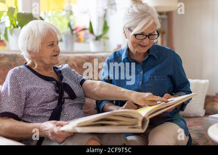 Ältere Frau und ihre Erwachsene Tochter betrachten Fotoalbum Zusammen auf der Couch im Wohnzimmer Stockfoto