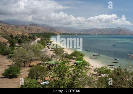 Von oben malerische Landschaft der Siedlung unter grünen Bäumen auf Seeufer mit Booten, die auf flachem Wasser gegen erstaunlich festgemacht sind highland Küste unter blauem Himmel mit üppigen weißen Wolken in Osttimor Stockfoto