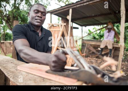 Uganda - 26. November 2016: Niedriger Winkel des fleißigen aufmerksamen afrikanischen Zimmermanns mit Handfläche während der Arbeit mit Holz in der Freiluft-rustikalen Werkstatt gegen ruhige verschwommene Mitarbeiter, die tagsüber auf einer Holzbank unter einem Metalldach ruhen Stockfoto