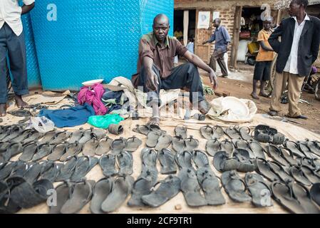 Uganda - 26. November 2016: Ruhiger nachdenklicher afrikanischer Rüde in legerer Kleidung, der Kamera anschaut, während er neben bunten Lumpen und Werkzeugen auf dem Boden sitzt und schmutzige schwarze Hausschuhe auf dem lokalen Markt gegen kleine rustikale Geschäfte verkauft Stockfoto