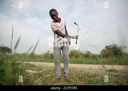 Uganda - November, 26 2016: Afrikanischer männlicher Teenager-Junge mit Axt, der an den Straßenschneideanlagen arbeitet Stockfoto