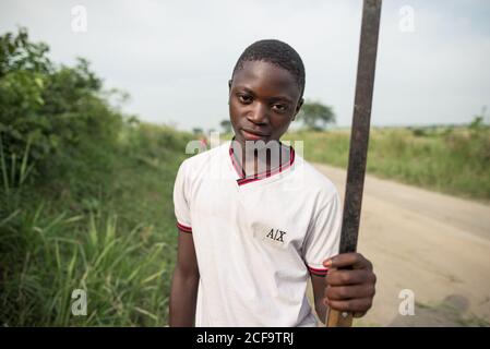 Uganda - November, 26 2016: Afrikanischer männlicher Teenager-Junge mit Axt, der auf der Straße arbeitet und Pflanzen schneidet und dabei die Kamera anschaut Stockfoto