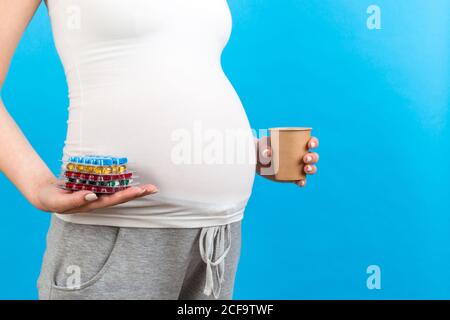 Nahaufnahme einer schwangeren Frau mit einem Papierbecher und einem Haufen Blasen mit Pillen auf buntem Hintergrund mit Kopieplatz. Behandlungskonzept. Stockfoto